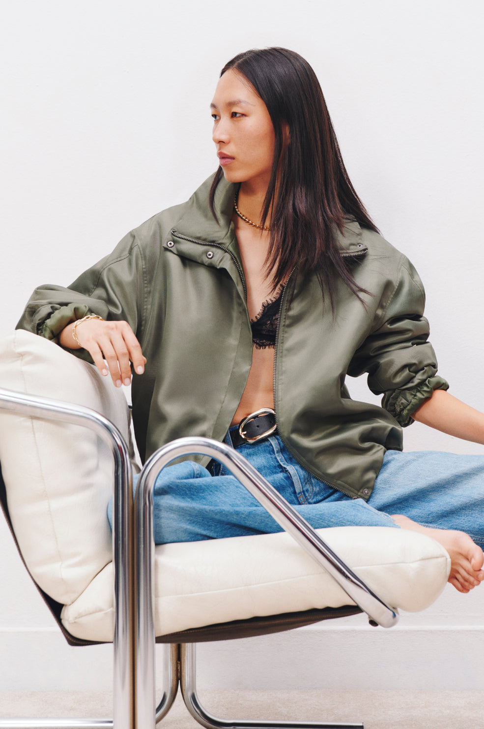 Woman sitting on a white chair wearing a green jacket and blue jeans.