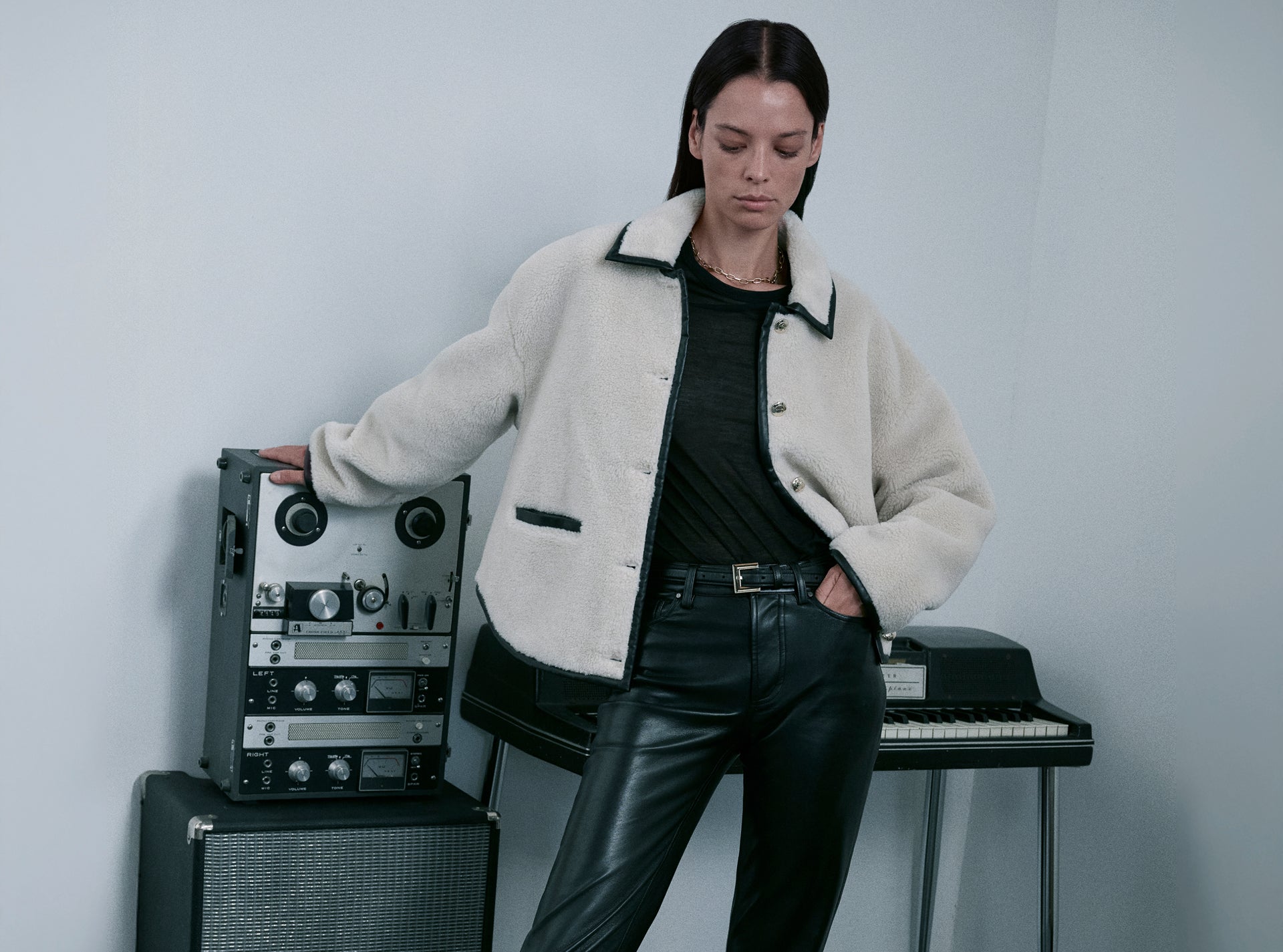 Woman in a white jacket and black pants standing next to electronic equipment against a plain background