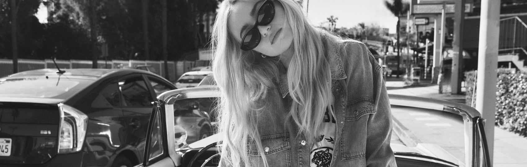 Person with long hair and sunglasses standing next to a car in a parking lot.