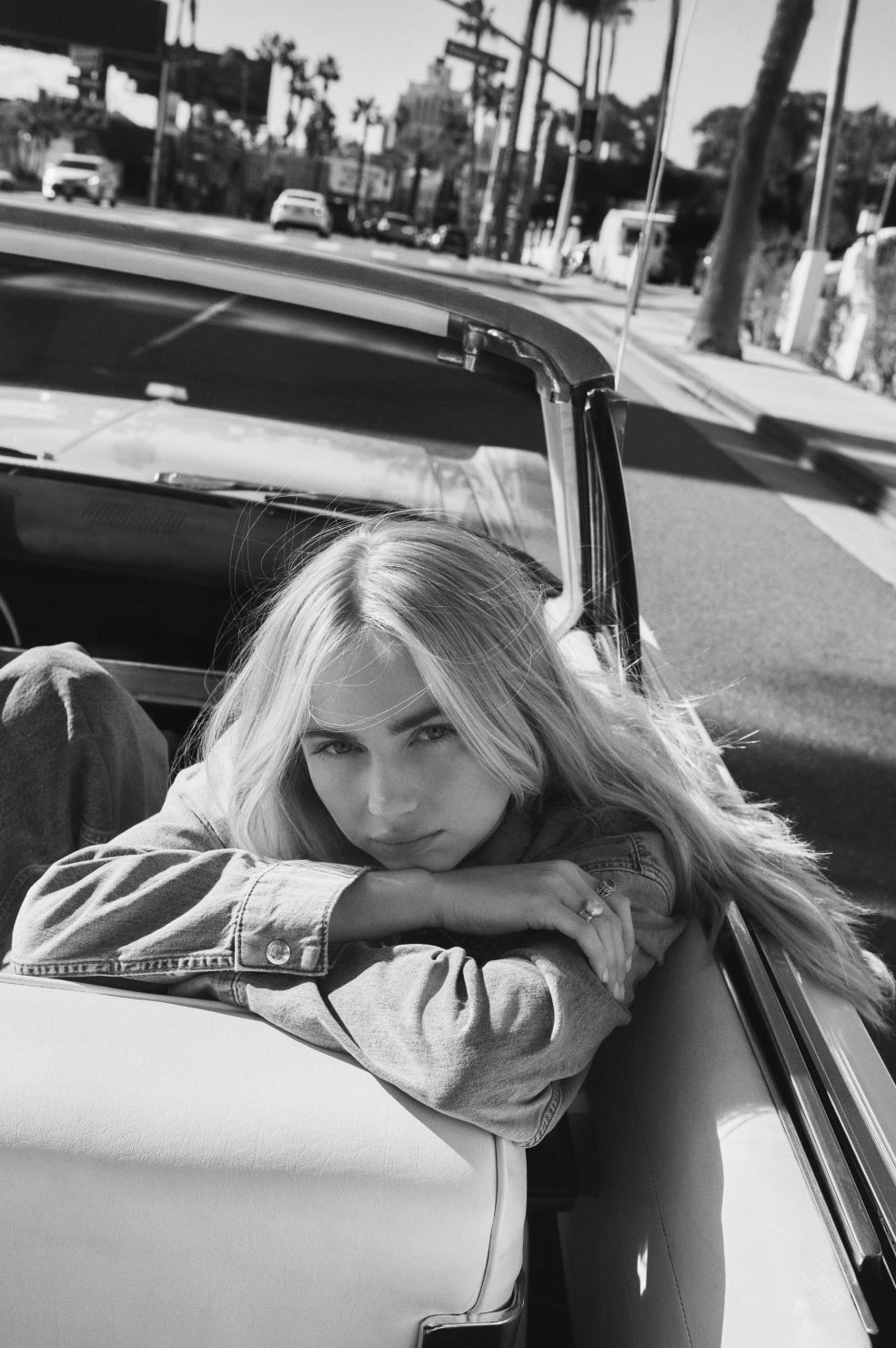 Person with long blonde hair leaning on a car door in a city street.
