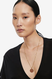 Model wearing a gold necklace with a circular charm, paired with a black v-neck top.